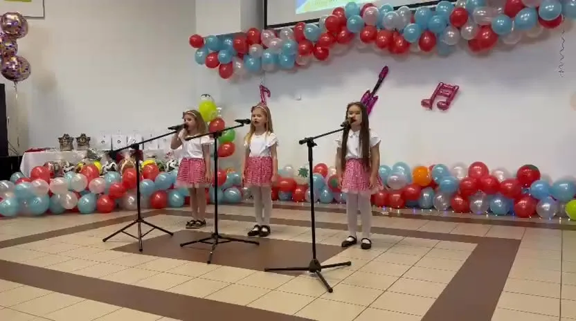 Trzy dziewczynki w białych bluzkach i czerwonych spódniczkach śpiewają do mikrofonów na tle balonów