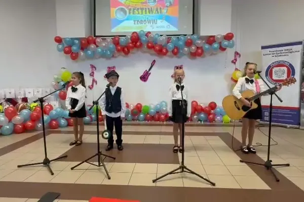 Czwórka uczniów w eleganckich strojach występuje, jedna z dziewczynek gra na gitarze