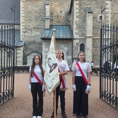 Poczet sztandarowy: troje uczniów w białych koszulach z biało-czerwonymi szarfami trzyma sztandar szkoły przed bramą kościoła
